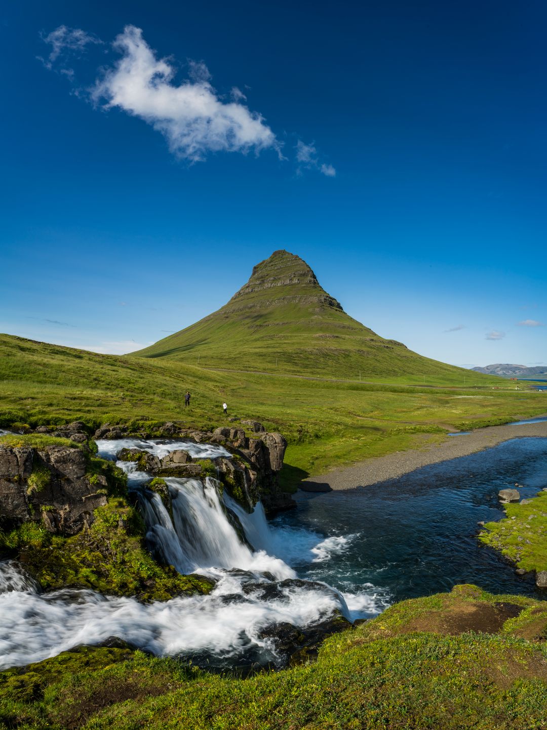 Kirkjufell with a rich blue sky and deep green grass with a waterfall and 1 cloud