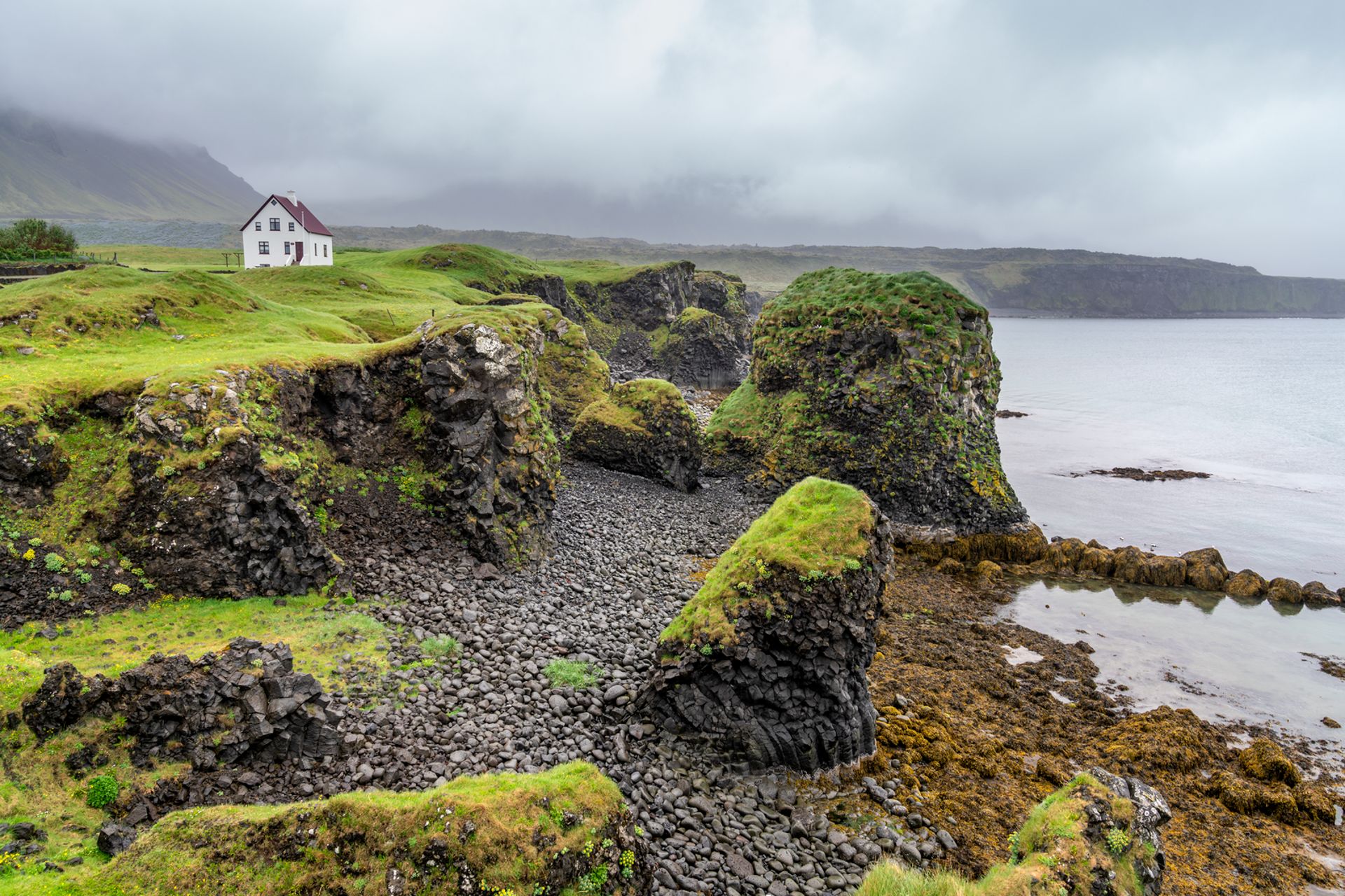 A cliff off of the coast with a small house on the edge and moss covered cliffs against black lava rock along the coast
