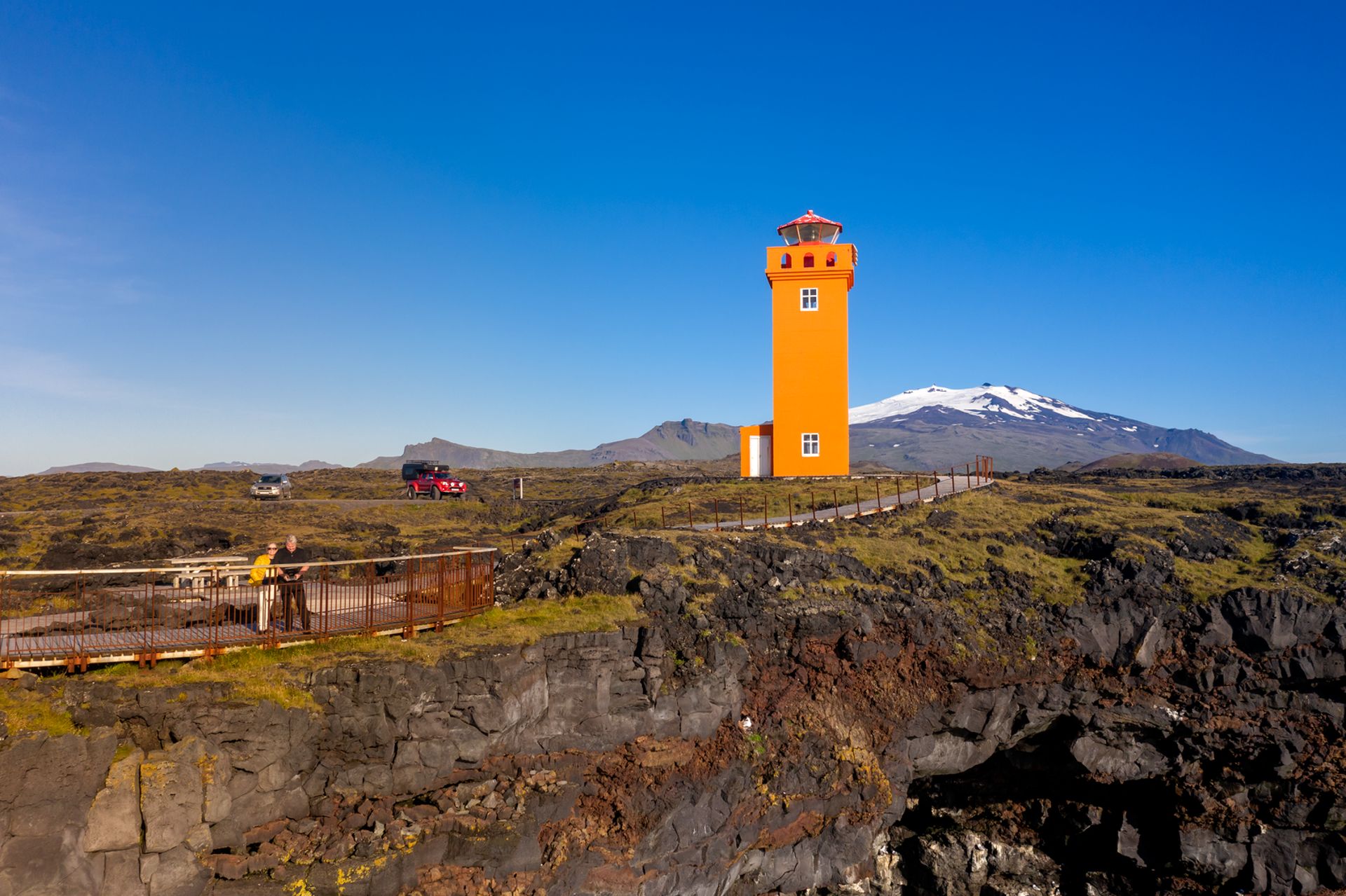 A bright orange light house standing in the middle of a moss covered lava field with a glacier topped mountain in the background