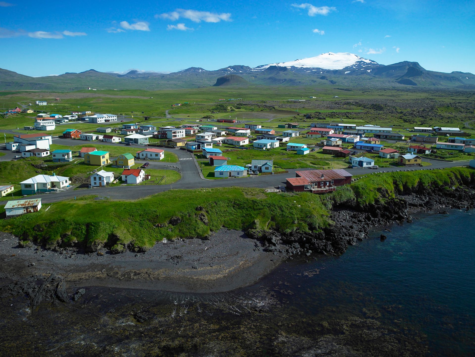 an aerial view of a small village off the coast of a blue ocean with a large ice capped glacier in the background on a blue sky day