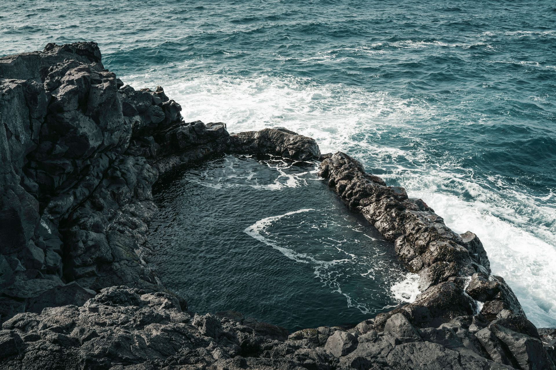 aerial view of a large lava rock pool off the coast of iceland by white cap waves crashing on the shore