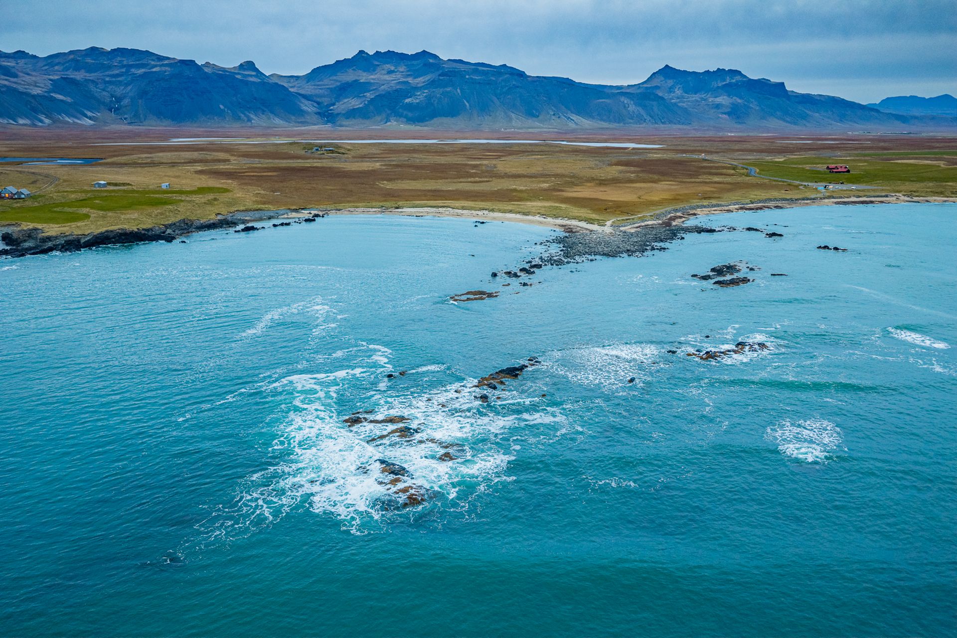 a clear blue ocean coast off of a small white sand beach in iceland with large mountains in the background.